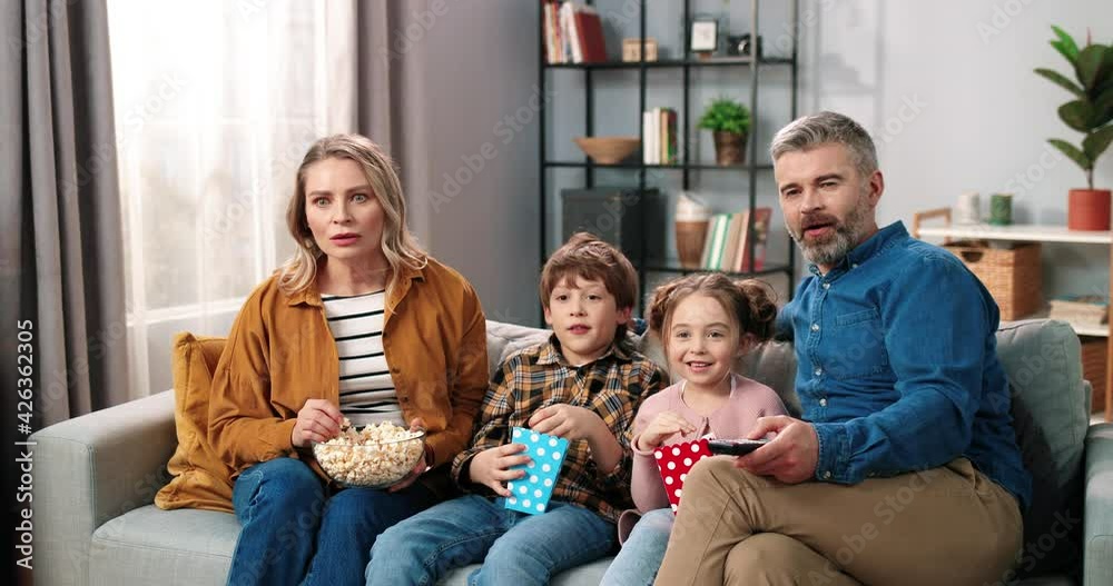 Happy family with two children sitting at home on couch and enthusiastically watching interesting movie eating popcorn and talking. Happy family with two children sitting at home on couch and enthusiastically watching interesting movie eating popcorn and talking.