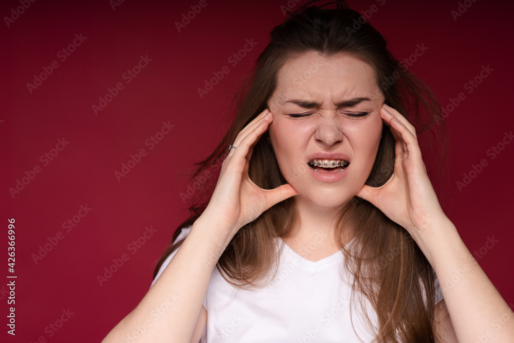 Beautiful woman grimaces and squints from pain in her teeth due to braces touching her temples with her hands