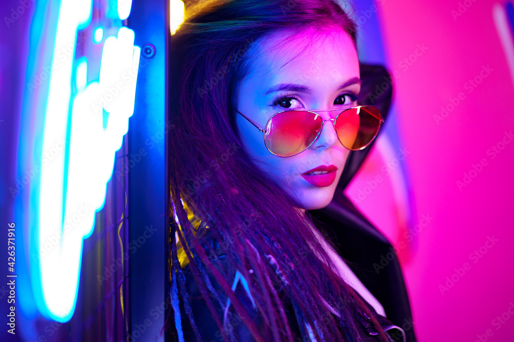 Fashion portrait of a young woman in sunglasses posing near neon signs in night club Fashion portrait of a young woman in sunglasses posing near neon signs in night club