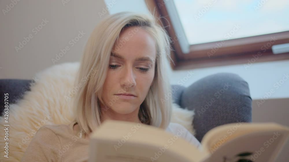 Slow motion of beautiful young woman reading book relaxing on armchair at home enjoying leisure time in solitude. People, lifestyle and hobby concept.