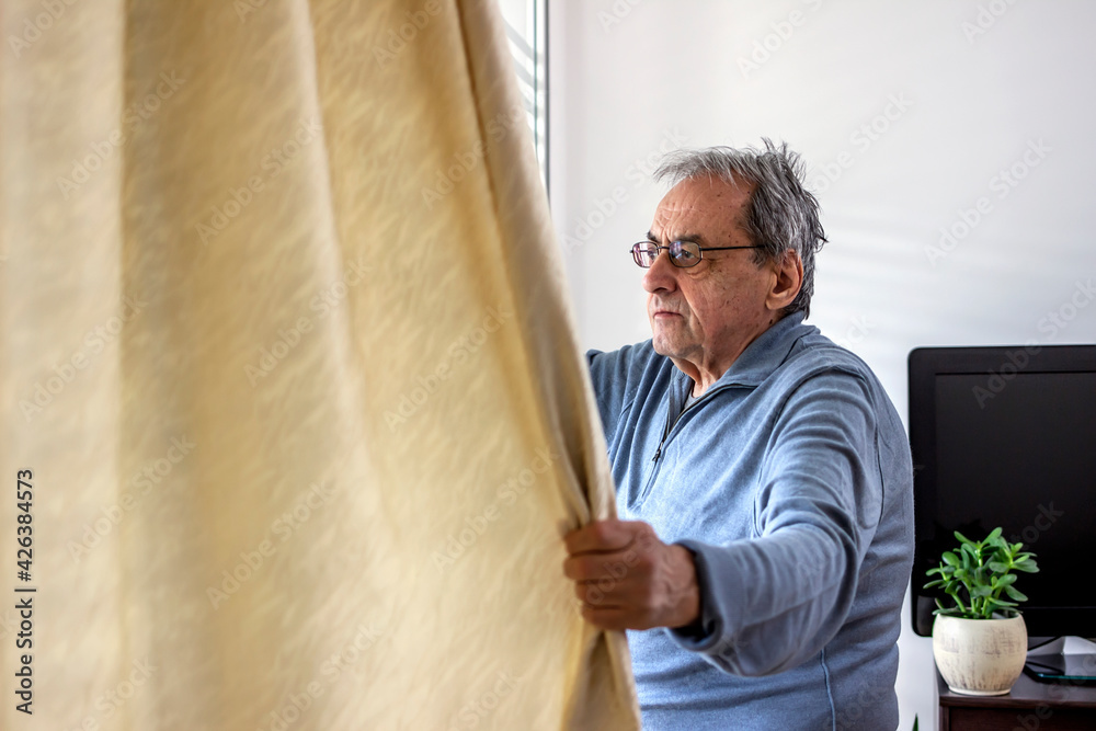 Fototapeta premium Elderly man staying home for safety during coronavirus pandemic and observing empty city through the window. Man staying home during coronavirus pandemic is looking through the window.