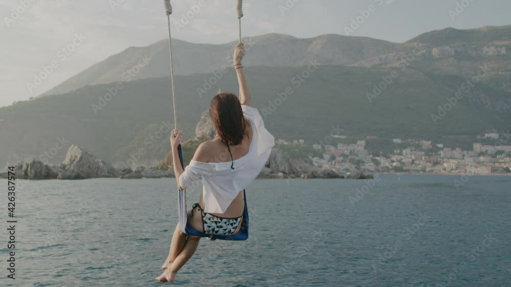 Back view brunette woman in a swimsuit sits on a swing above the sea ...