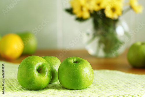 green fresh apples in the kitchen, just washed lie on a towel dry, water drops on the fruit, in the background lemon and a vase with yellow flowers blurred