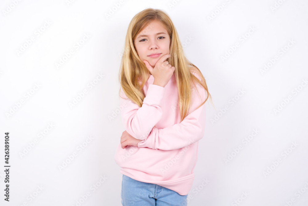 Thoughtful smiling beautiful caucasian little girl wearing pink hoodie over white background keeps hand under chin, looks directly at camera, listens something with interest. Youth concept.