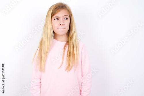 Amazed puzzled beautiful caucasian little girl wearing pink hoodie over white background  curves lips and has worried look  sees something awful in front.