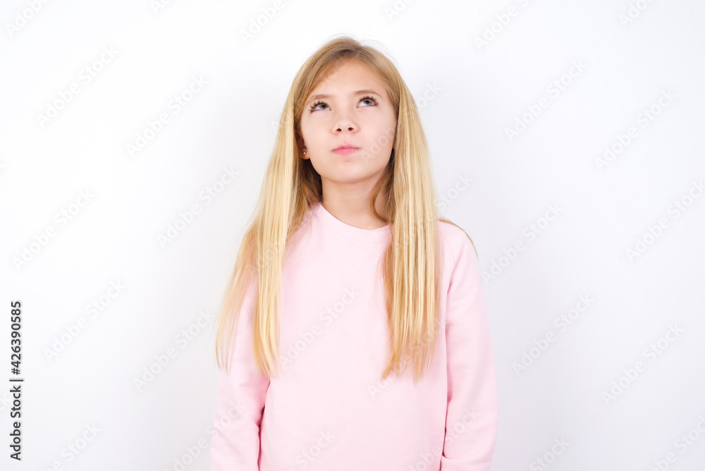 beautiful caucasian little girl wearing pink hoodie over white background has worried face looking up lips together, being upset thinking about something important, keeps hands down. beautiful caucasian little girl wearing pink hoodie over white background has worried face looking up lips together, being upset thinking about something important, keeps hands down.