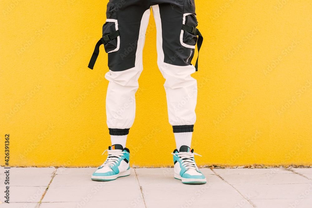 Naklejka premium Unrecognizable young trendy and fashionable man against a yellow wall wearing a black and white pants with basket sneakers