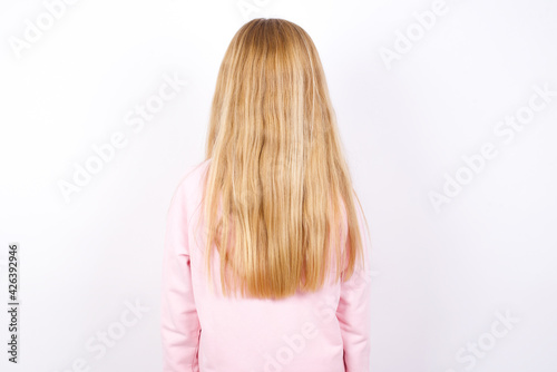 beautiful caucasian little girl wearing pink hoodie over white background standing backwards looking away with arms on body.