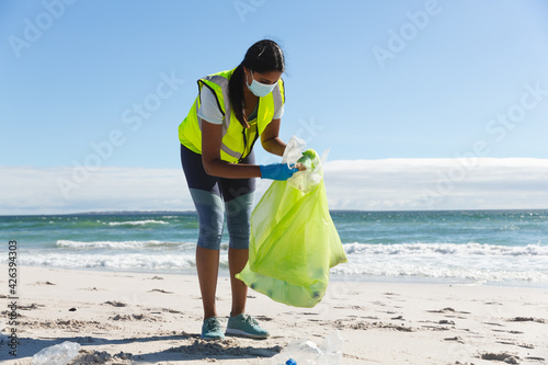 Mixed race woman wearing face mask collecting rubbish from the beach