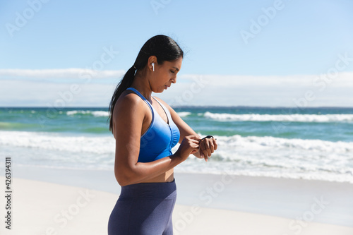 Mixed race woman exercising on beach wearing wireless earphones using smartwatch