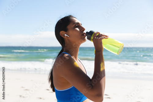 Mixed race woman exercising on beach wearing wireless earphones drinking water