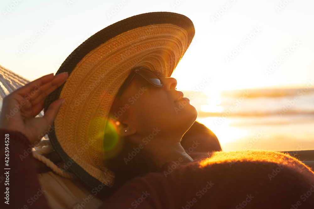 Mixed race woman on beach holiday lying in hammock during sunset - obrazy, fototapety, plakaty 