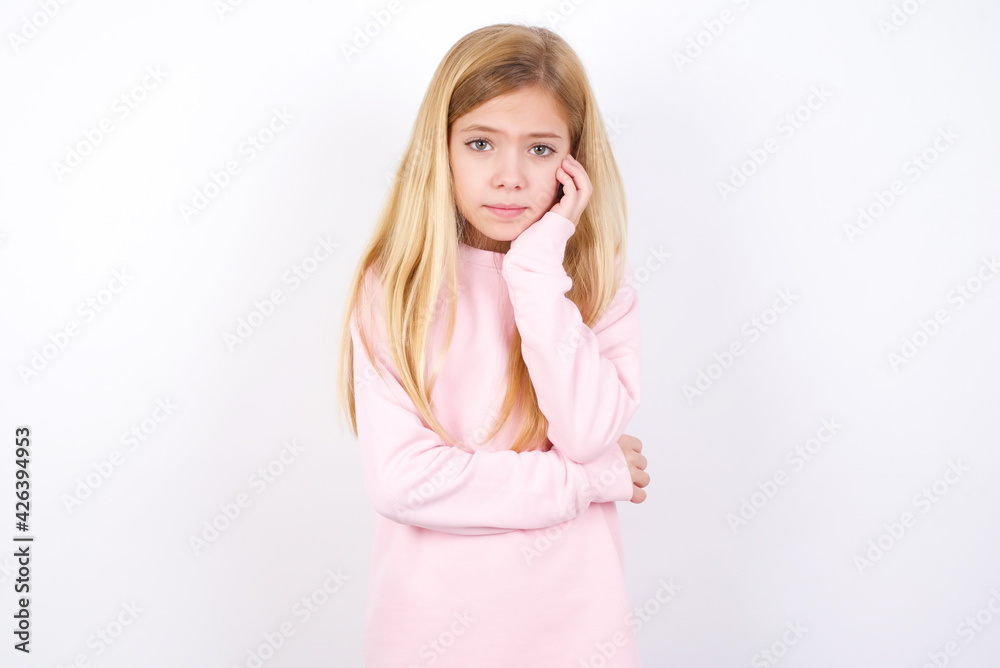Very bored beautiful caucasian little girl wearing pink hoodie over white background holding hand on cheek while support it with another crossed hand, looking tired and sick. Very bored beautiful caucasian little girl wearing pink hoodie over white background holding hand on cheek while support it with another crossed hand, looking tired and sick.