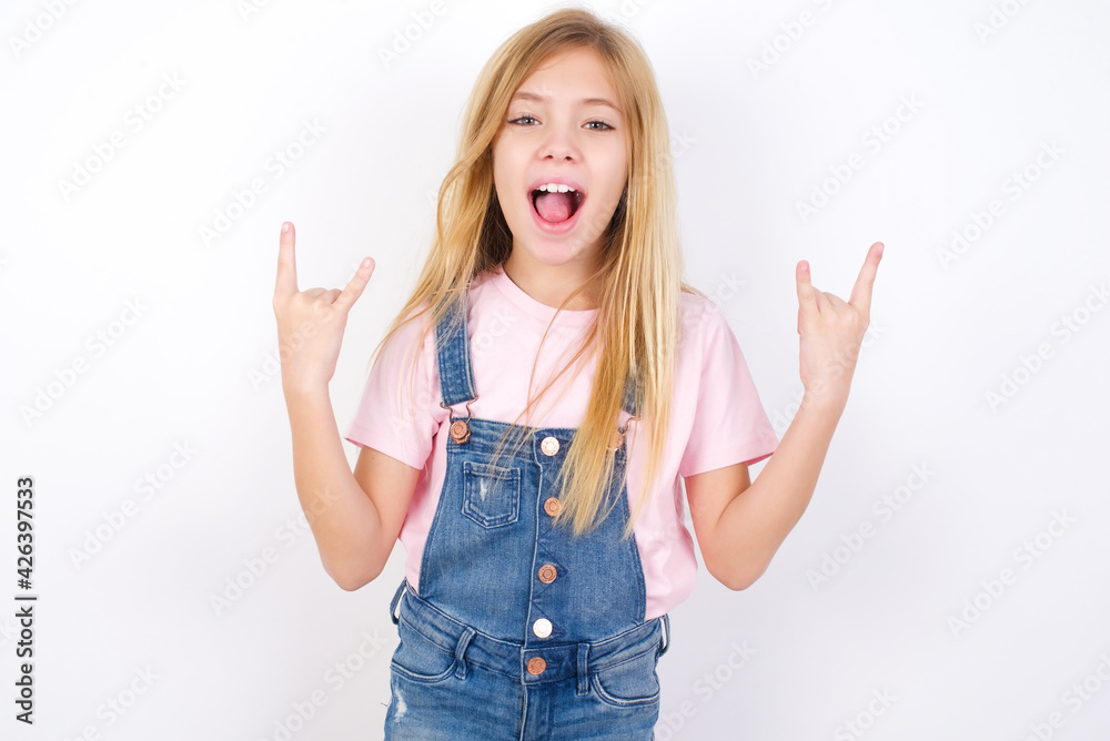 beautiful Caucasian little girl wearing denim overalls over white background makes rock n roll sign looks self confident and cheerful enjoys cool music at party. Body language concept. beautiful Caucasian little girl wearing denim overalls over white background makes rock n roll sign looks self confident and cheerful enjoys cool music at party. Body language concept.