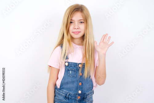 beautiful Caucasian little girl wearing denim overalls over white background shows middle finger bad sign asks not to bother. Provocation and rude attitude.