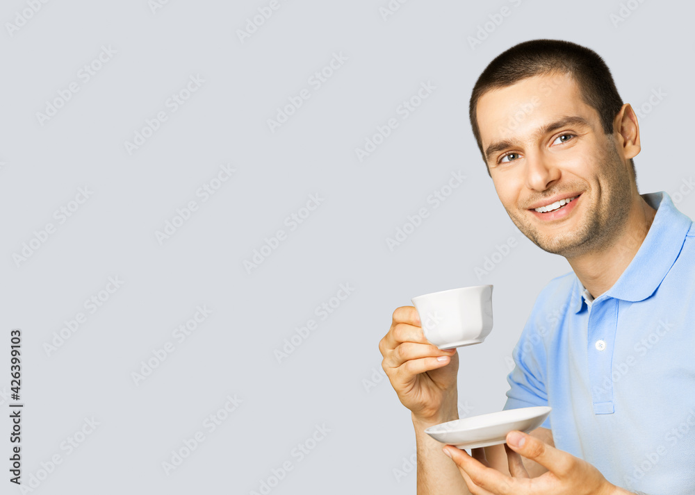 Portrait of young happy smiling man in blue casual clothing drinking coffee, isolated on grey color background, with copy space blank area for some slogan or text. Portrait of young happy smiling man in blue casual clothing drinking coffee, isolated on grey color background, with copy space blank area for some slogan or text.