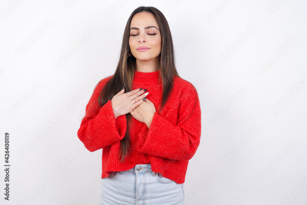 young beautiful brunette woman wearing red sweater closes eyes and keeps hands on chest near heart, expresses sincere emotions, being kind hearted and honest. Body language and real feelings concept. young beautiful brunette woman wearing red sweater closes eyes and keeps hands on chest near heart, expresses sincere emotions, being kind hearted and honest. Body language and real feelings concept.