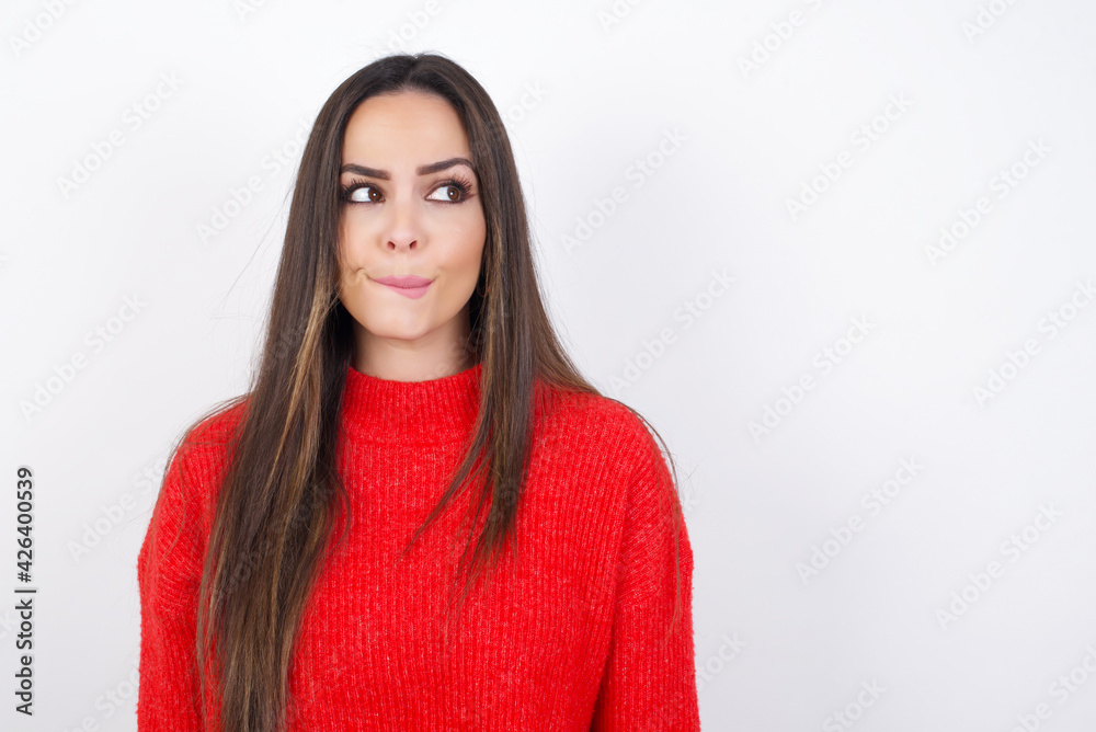 Amazed puzzled young beautiful brunette woman wearing red knitted sweater over white wall, curves lips and has worried look, sees something awful in front.