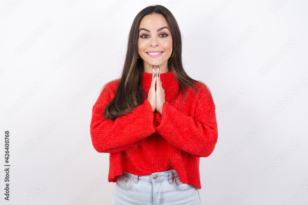 young beautiful brunette woman wearing red knitted sweater over white wall keeps palms together, has pleased expression. Glad attractive male makes request, pleads for mercy. Hopeful young adult. young beautiful brunette woman wearing red knitted sweater over white wall keeps palms together, has pleased expression. Glad attractive male makes request, pleads for mercy. Hopeful young adult.