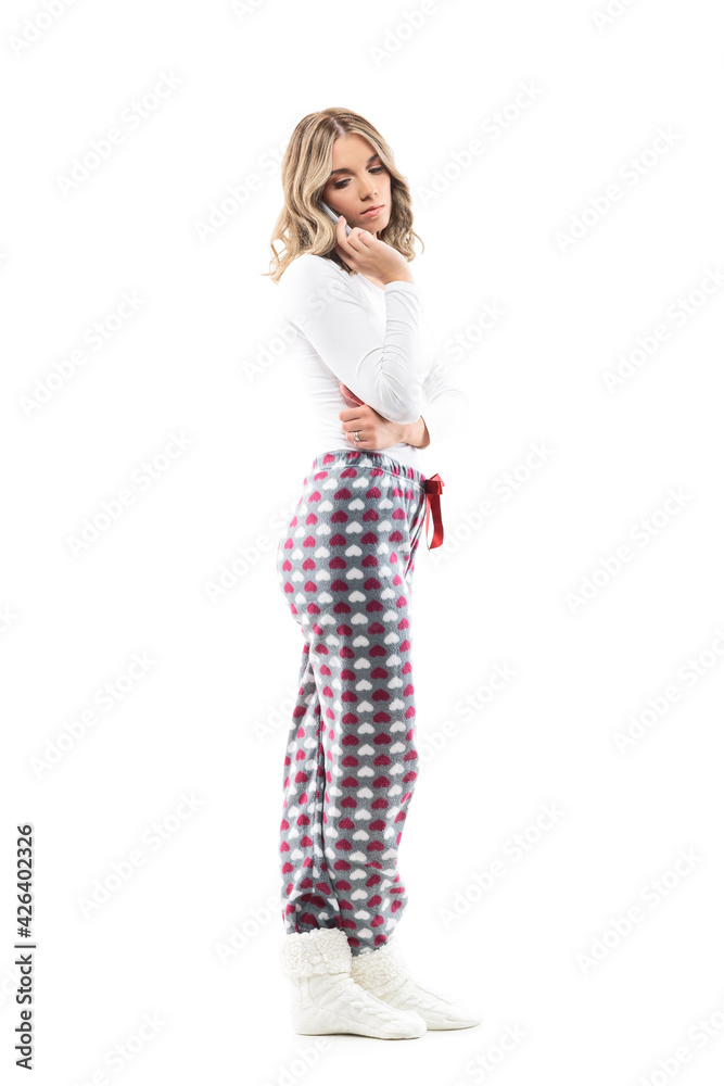 Emotional pretty young woman in pajamas talking on the cell phone looking down over shoulder. Full body length isolated on white background. Emotional pretty young woman in pajamas talking on the cell phone looking down over shoulder. Full body length isolated on white background.