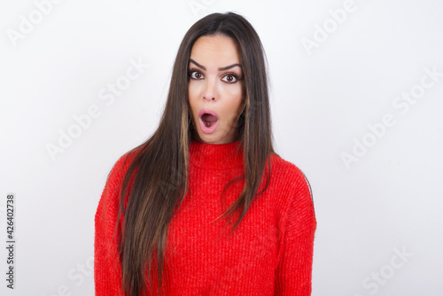 Expressive facial expressions. Shocked stupefied young beautiful brunette woman wearing red knitted sweater over white wall, keeps jaw dropped feels stunned from what he sees aside. © Jihan