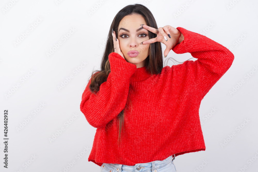 young beautiful brunette woman wearing red knitted sweater over white wall making v-sign near eyes. Leisure lifestyle people person celebrate flirt coquettish concept. young beautiful brunette woman wearing red knitted sweater over white wall making v-sign near eyes. Leisure lifestyle people person celebrate flirt coquettish concept.