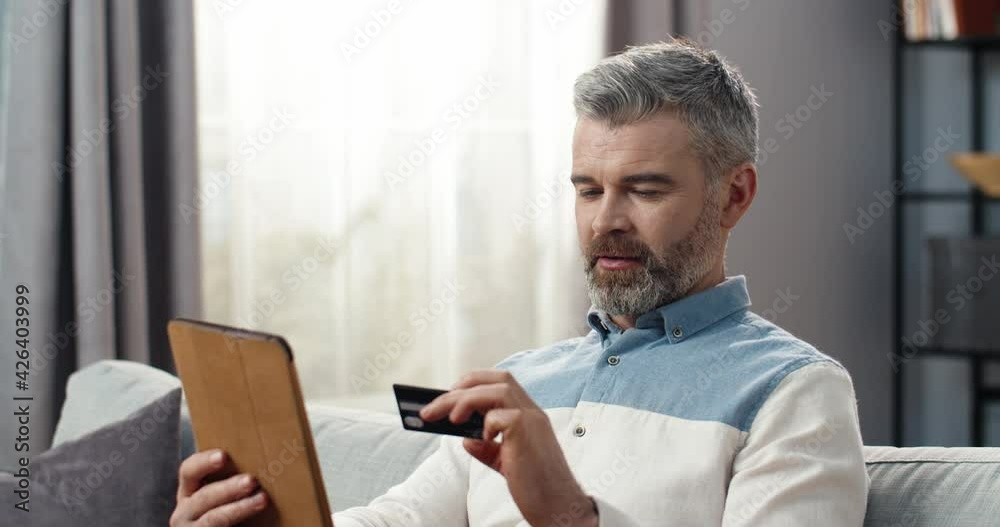 Happy adult man sitting at home on couch holding tablet enters bank data from the card and rejoices in successful purchase. Happy adult man sitting at home on couch holding tablet enters bank data from the card and rejoices in successful purchase.