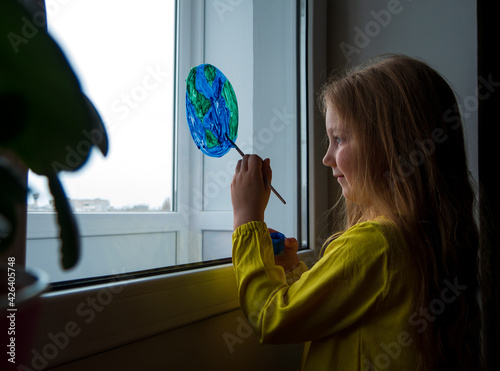 Cute little girl painting planet on window at home. Happy Earth Day April 22 greeting message. Creative family leisure lockdown new reality. Ecology Saving environment conscious consumption concept. photo