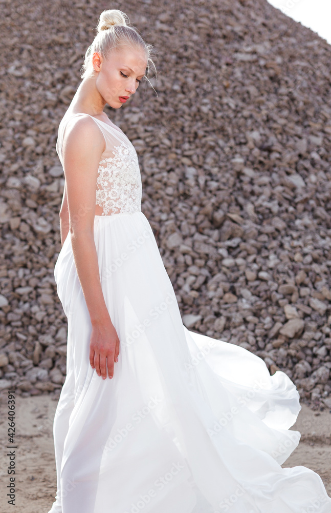 Profile view of the blonde young woman in a white wedding dress flying in wind, posed behind piles of stones. Profile view of the blonde young woman in a white wedding dress flying in wind, posed behind piles of stones.