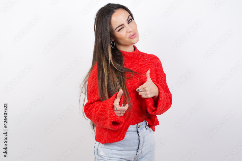 Hey you, bang. Joyful and charismatic good-looking young beautiful brunette woman wearing red knitted sweater over white wall winking and pointing with finger pistols at camera happily and cheeky. Hey you, bang. Joyful and charismatic good-looking young beautiful brunette woman wearing red knitted sweater over white wall winking and pointing with finger pistols at camera happily and cheeky.