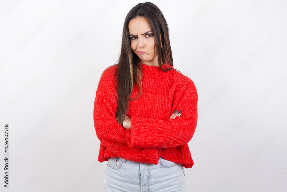 Picture of angry young beautiful brunette woman wearing red knitted sweater over white wall crossing arms. Looking at camera with disappointed expression. Picture of angry young beautiful brunette woman wearing red knitted sweater over white wall crossing arms. Looking at camera with disappointed expression.