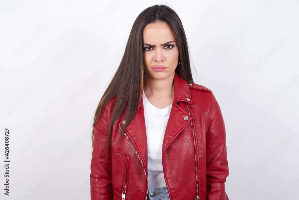 Offended dissatisfied young beautiful brunette woman wearing red biker jacket over white wall with moody displeased expression at camera being disappointed by something Offended dissatisfied young beautiful brunette woman wearing red biker jacket over white wall with moody displeased expression at camera being disappointed by something