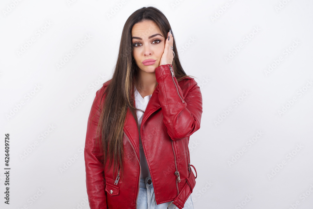 Sad lonely young beautiful brunette woman wearing red biker jacket over white wall touches cheek with hand bites lower lip and gazes with displeasure. Bad emotions Sad lonely young beautiful brunette woman wearing red biker jacket over white wall touches cheek with hand bites lower lip and gazes with displeasure. Bad emotions