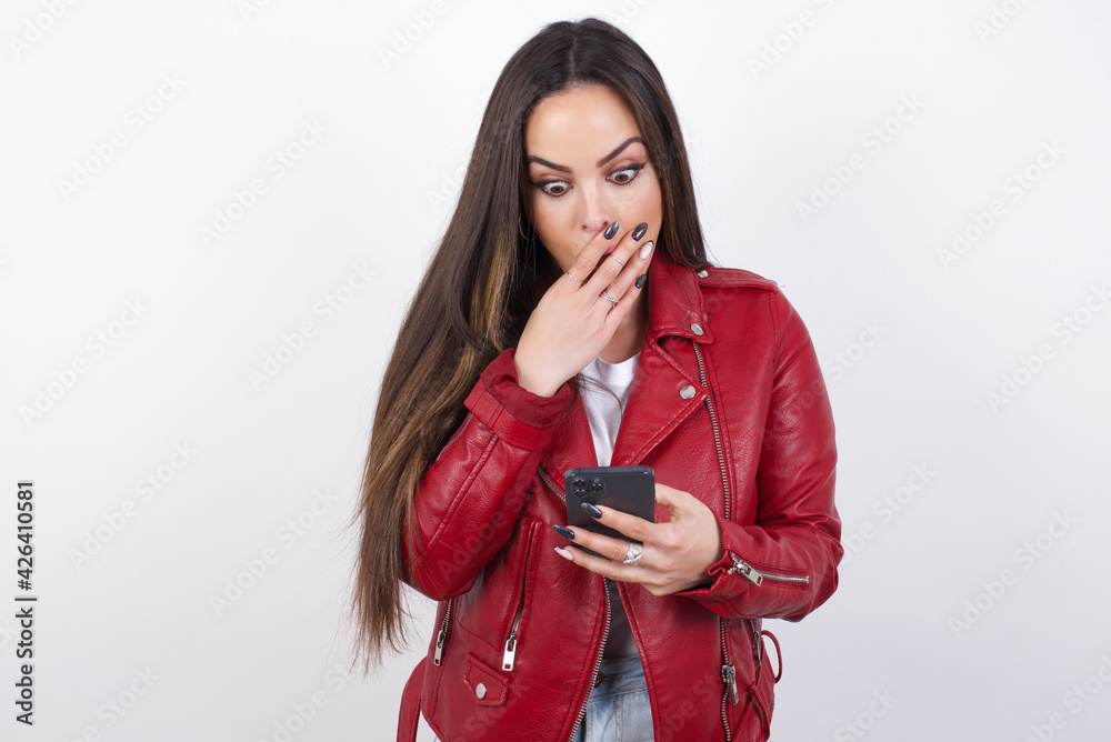 Naklejka premium young beautiful brunette woman wearing red biker jacket over white wall being deeply surprised, stares at smartphone display, reads shocking news on website, Omg, its horrible!