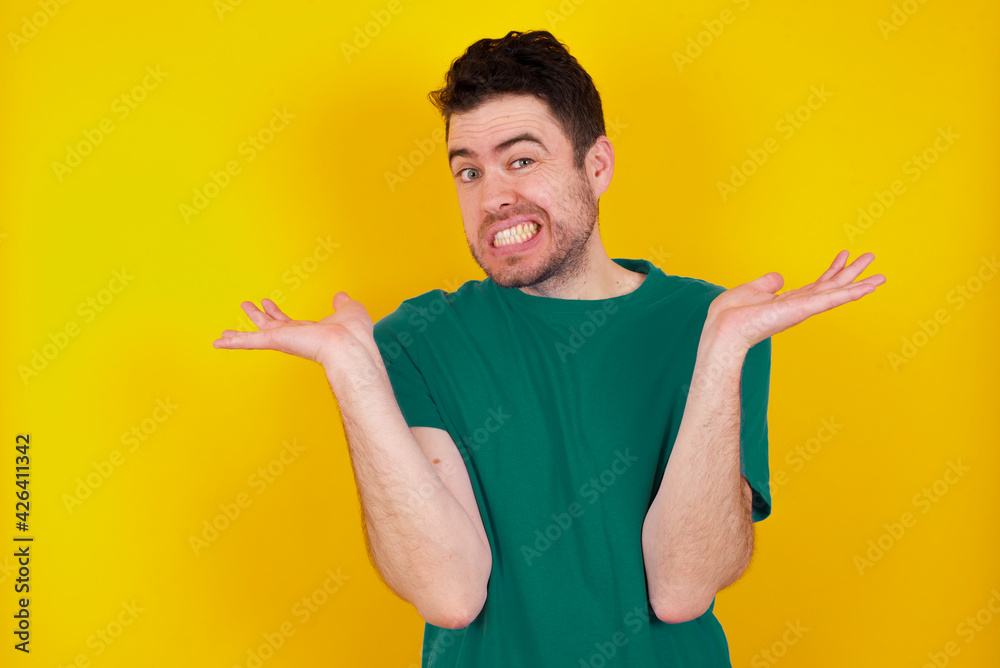 Clueless young Caucasian man wearing green T-shirt against yellow wall shrugs shoulders with hesitation, faces doubtful situation, spreads palms, Hard decision Clueless young Caucasian man wearing green T-shirt against yellow wall shrugs shoulders with hesitation, faces doubtful situation, spreads palms, Hard decision