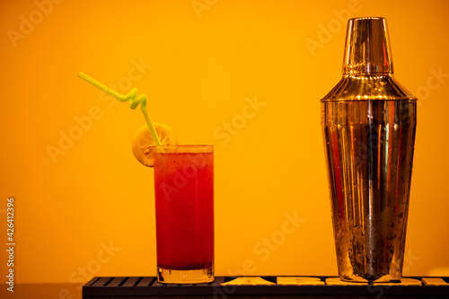 Red cocktail in a glass and a cocktail shaker next to it with an orange background 