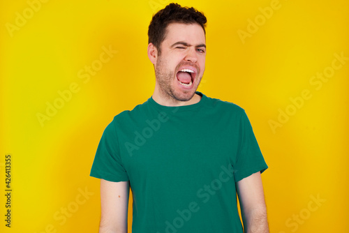 young Caucasian man wearing green T-shirt against yellow wall winking looking at the camera with sexy expression, cheerful and happy face.