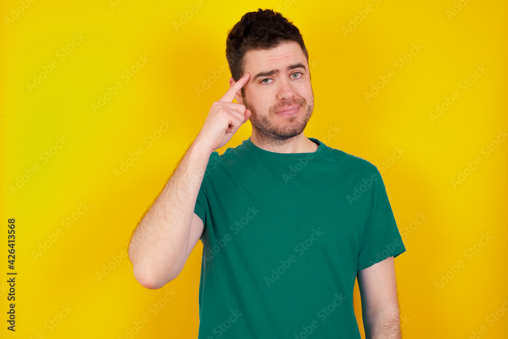 young Caucasian man wearing green T-shirt against yellow wall tries to memorize something, keeps fore finger on temple, reminds information for exam, young Caucasian man wearing green T-shirt against yellow wall tries to memorize something, keeps fore finger on temple, reminds information for exam,
