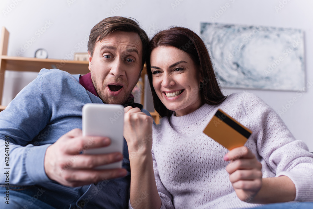 Excited couple with credit card using smartphone on blurred foreground