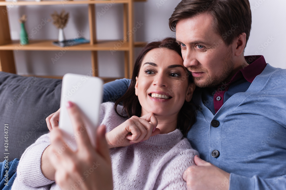 Positive woman holding cellphone on blurred foreground near husband in living room