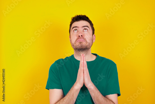 young Caucasian man wearing green T-shirt against yellow wall begging and praying with hands together with hope expression on face very emotional and worried. Please God