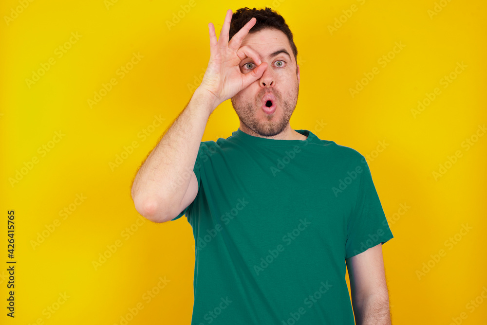 young Caucasian man wearing green T-shirt against yellow wall doing ok gesture shocked with surprised face, eye looking through fingers. Unbelieving expression. young Caucasian man wearing green T-shirt against yellow wall doing ok gesture shocked with surprised face, eye looking through fingers. Unbelieving expression.