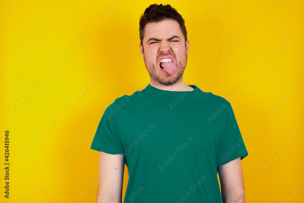 young Caucasian man wearing green T-shirt against yellow wall sticking tongue out happy with funny expression. Emotion concept. young Caucasian man wearing green T-shirt against yellow wall sticking tongue out happy with funny expression. Emotion concept.