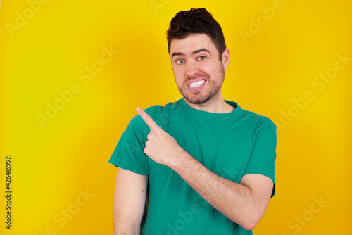 young Caucasian man wearing green T-shirt against yellow wall Pointing aside worried and nervous with forefinger, concern and surprise concept.