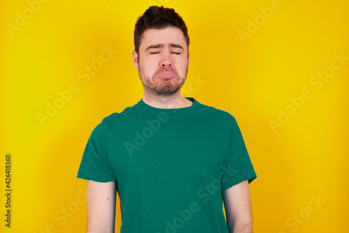 Dismal gloomy rejected young handsome caucasian man wearing green t-shirt against yellow background has problems and difficulties, curves lower lip and closes eyes in despair, being in depression