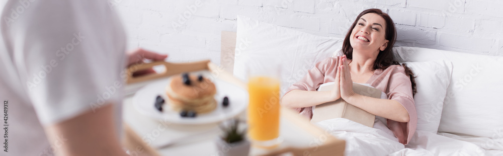 Cheerful woman with book showing praying hands near husband with breakfast on blurred foreground in bedroom, banner Cheerful woman with book showing praying hands near husband with breakfast on blurred foreground in bedroom, banner