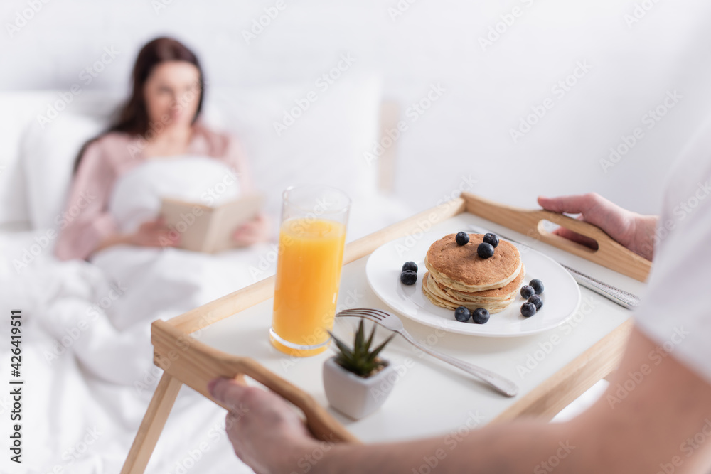 Man holding breakfast on tray near wife on blurred background in bedroom Man holding breakfast on tray near wife on blurred background in bedroom