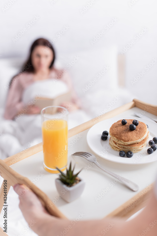 Man holding tray with pancakes and orange juice near wife on blurred background