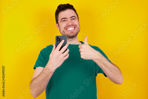 Portrait of young handsome caucasian man wearing green t-shirt against yellow background using and texting with smartphone happy with big smile doing ok sign, thumb up with fingers, excellent sign