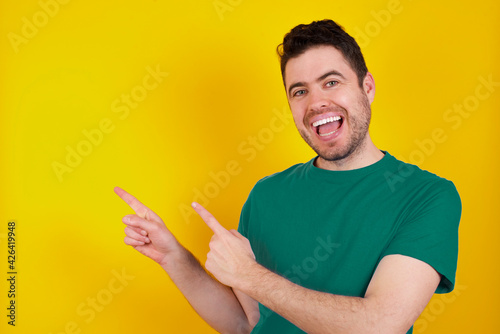 young handsome caucasian man wearing green t-shirt against yellow background point at copyspace recommend sales discounts